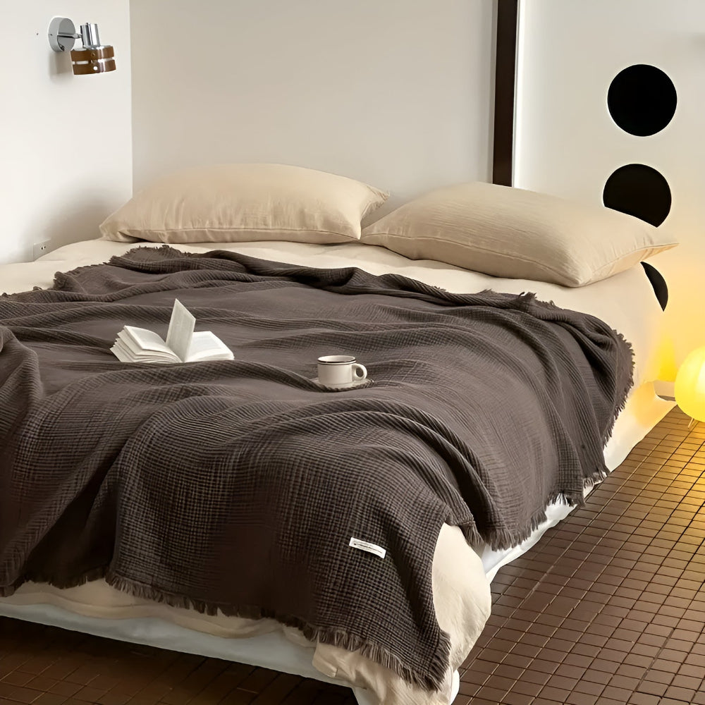 A dark brown cotton crinkle tassel blanket is draped over a bed with two light beige pillows.