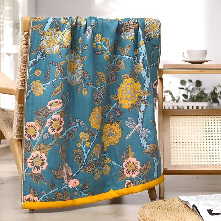 Teal throw blanket with a floral and bird pattern in yellow, pink, and gray, draped over a chair.
