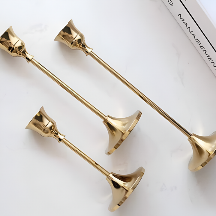 Three gold taper candle holders of varying sizes arranged on a white marble surface.