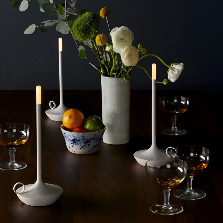 Portable aluminum table lamp with a warm glow on a wooden table with drinks and fruit.