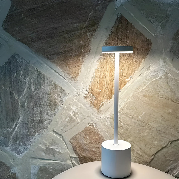 White cordless table lamp with a cylindrical base and a flat top light, casting a warm glow against a textured stone wall.