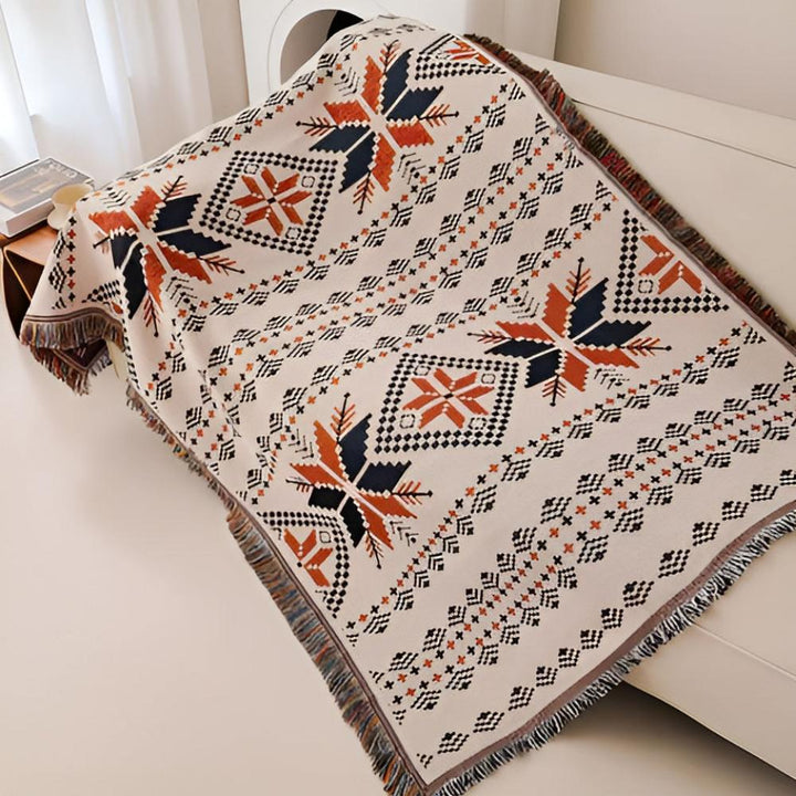 Cream throw blanket with a repeating geometric snowflake pattern in navy, orange, and black, featuring fringed edges.