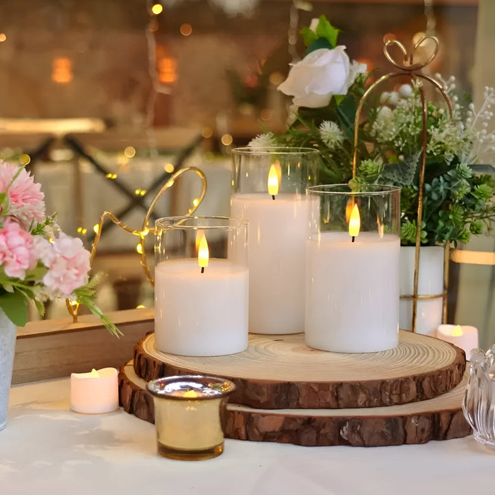 Three white flameless candles in glass cylinders on wood slices, with fairy lights and flowers.