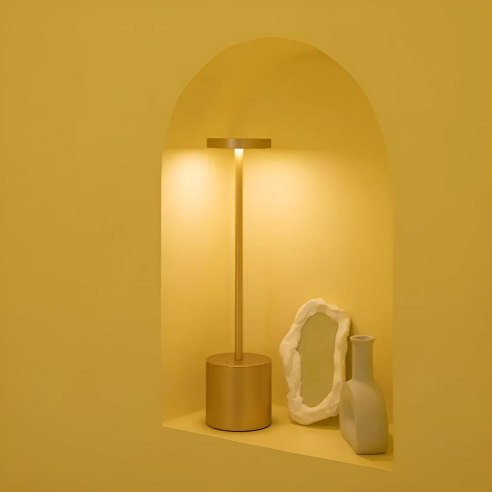 Gold cordless table lamp with a warm glow, placed on a shelf with a decorative mirror and vase.