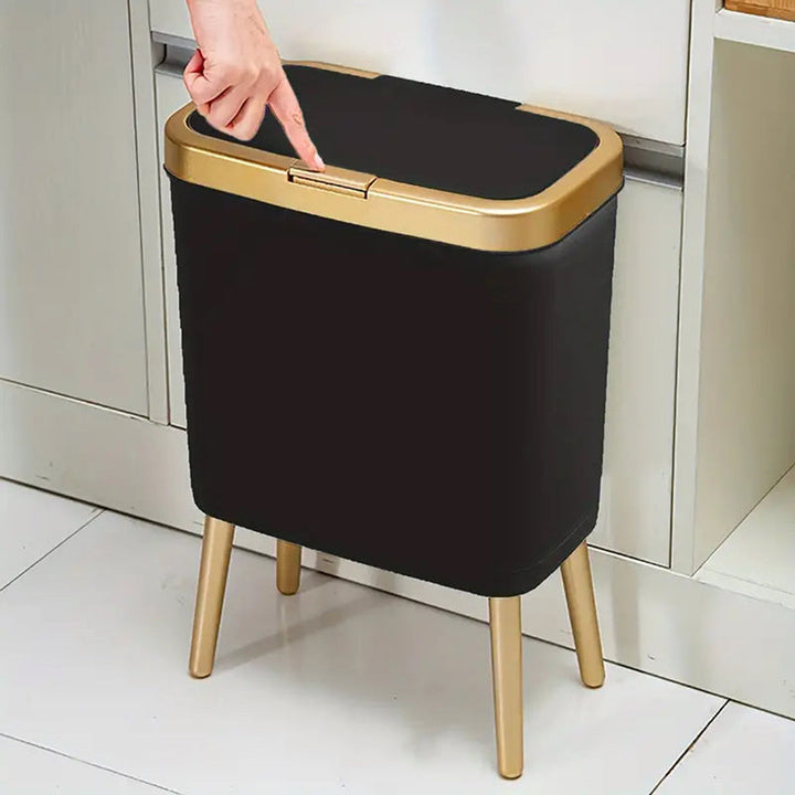 Black 15L elevated trash can with gold trim and legs, lid being opened by a finger.