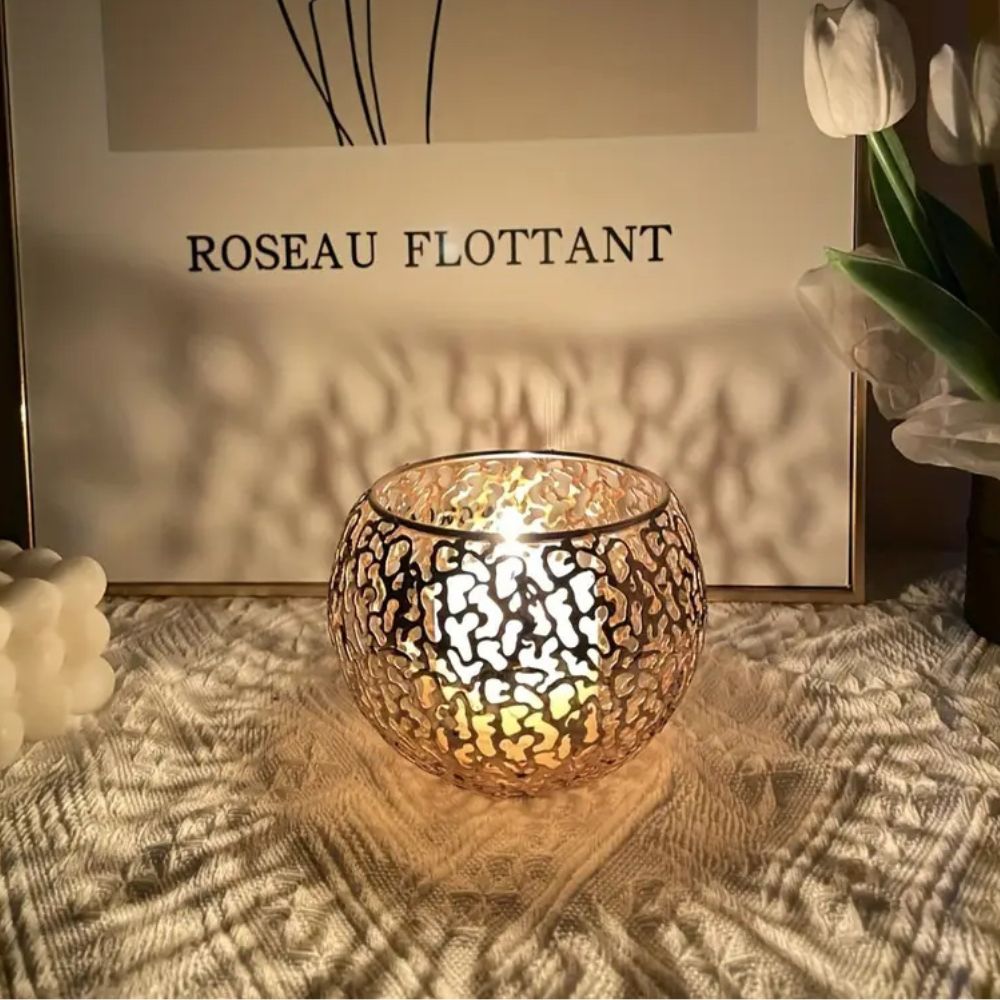 A lit tealight candle inside a round, metallic, geometric patterned holder, casting shadows.