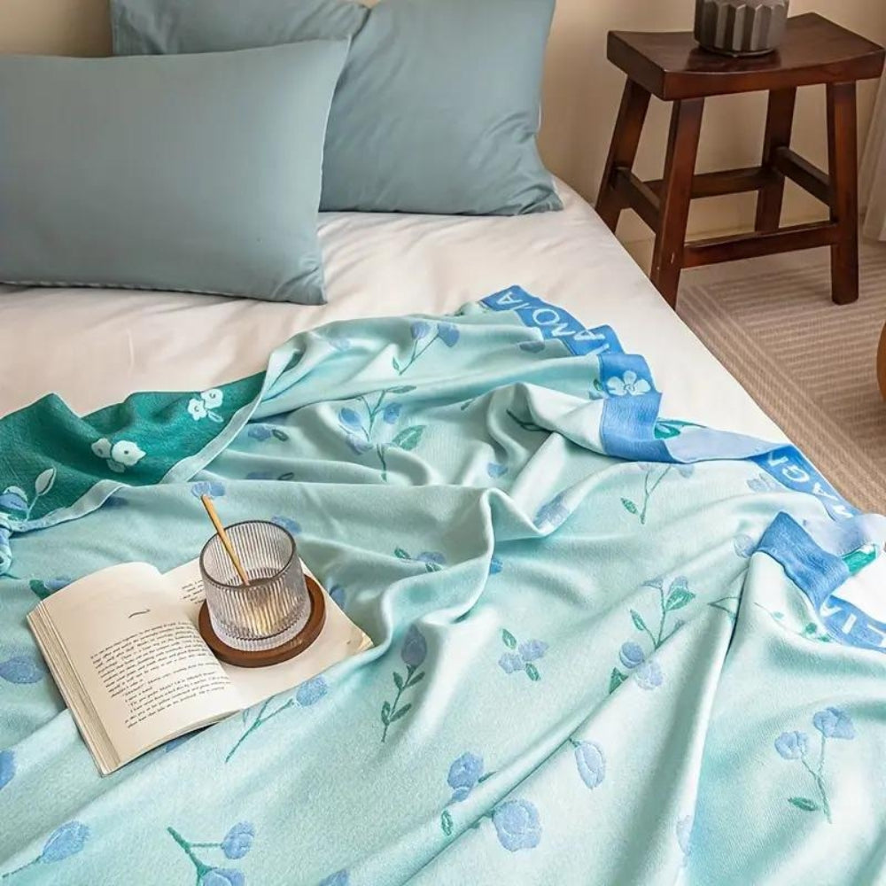 Light blue floral bamboo cooling blanket draped on a bed with a book and glass.