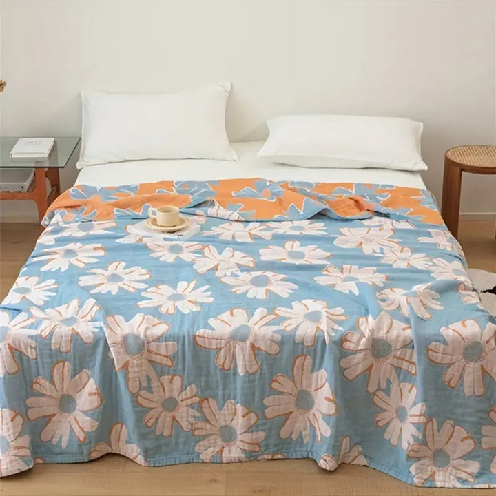 Blue cotton blanket with a large white and orange daisy pattern, draped on a bed with white pillows.