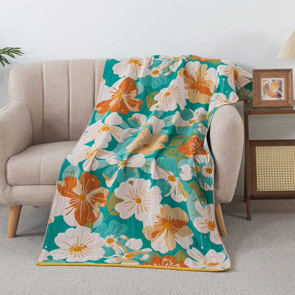 A teal cotton gauze blanket with a tropical floral pattern of orange, white, and yellow flowers draped over a cream sofa.