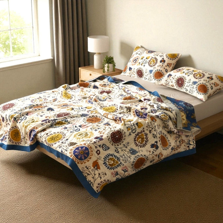 A bed with a white cotton gauze coverlet featuring a colorful floral and circular pattern. Two pillows with the same pattern are on the bed.