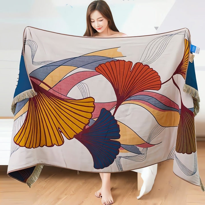 A woman holds a large, light-colored cotton gauze towel with a ginkgo leaf print and tassel edges.
