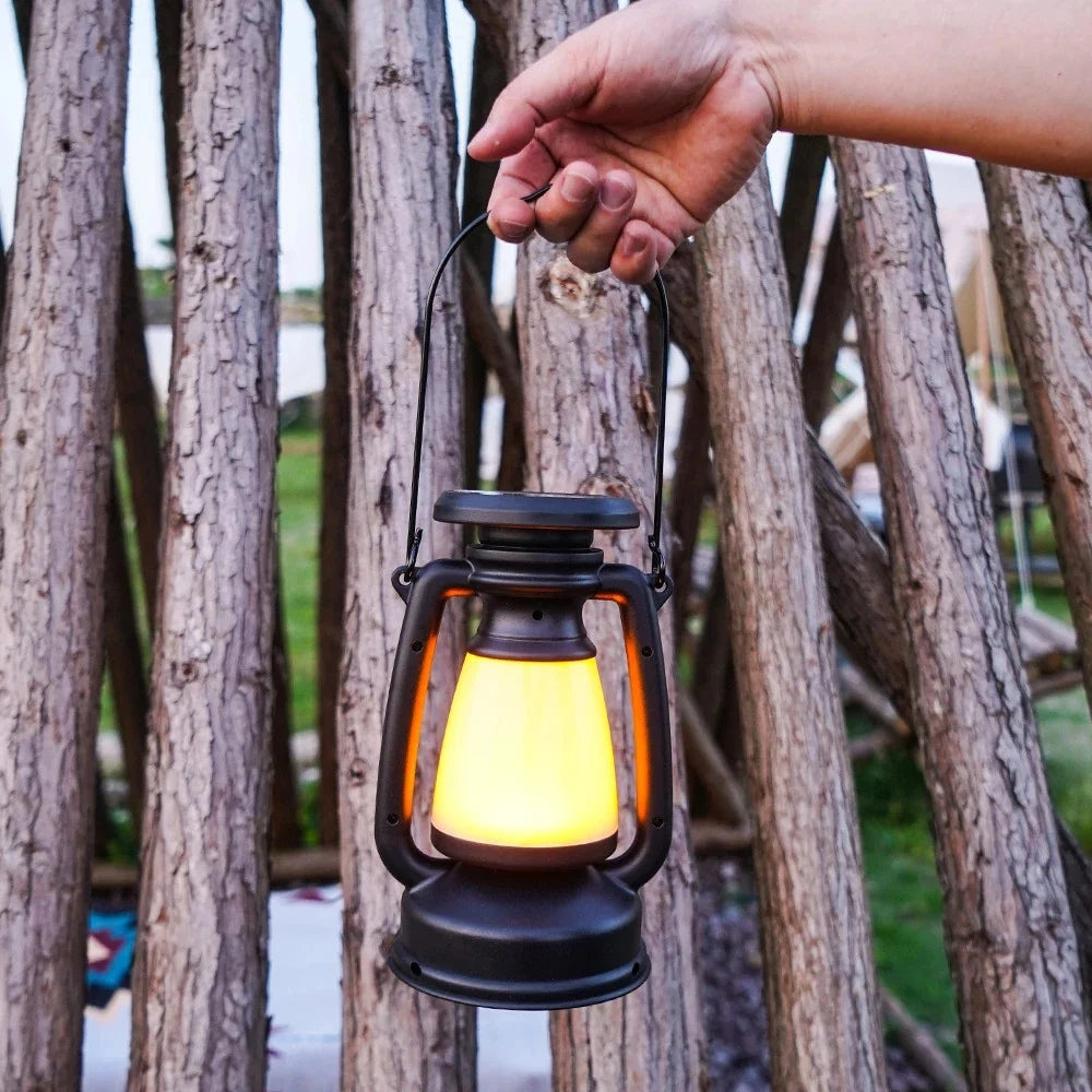 Black solar camping lantern with a warm yellow light, held by a hand against a backdrop of wooden logs.