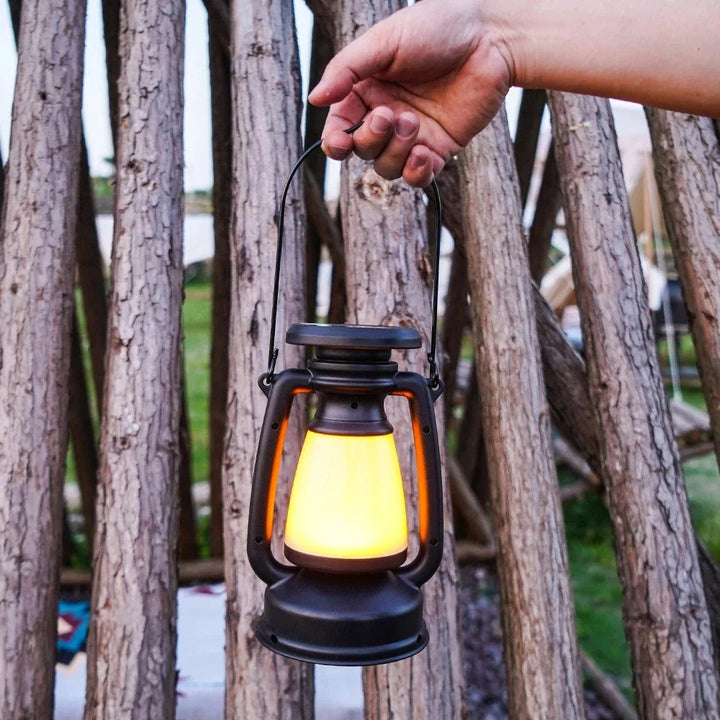 Black solar camping lantern with a warm yellow light, held by a hand against a backdrop of wooden logs.