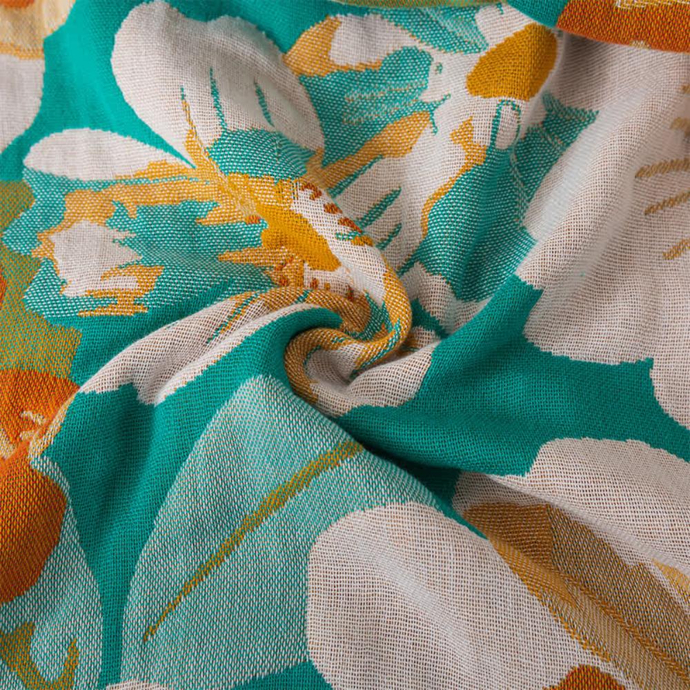 A close-up of a teal cotton gauze bath towel with a bold floral pattern in white, yellow, and orange.