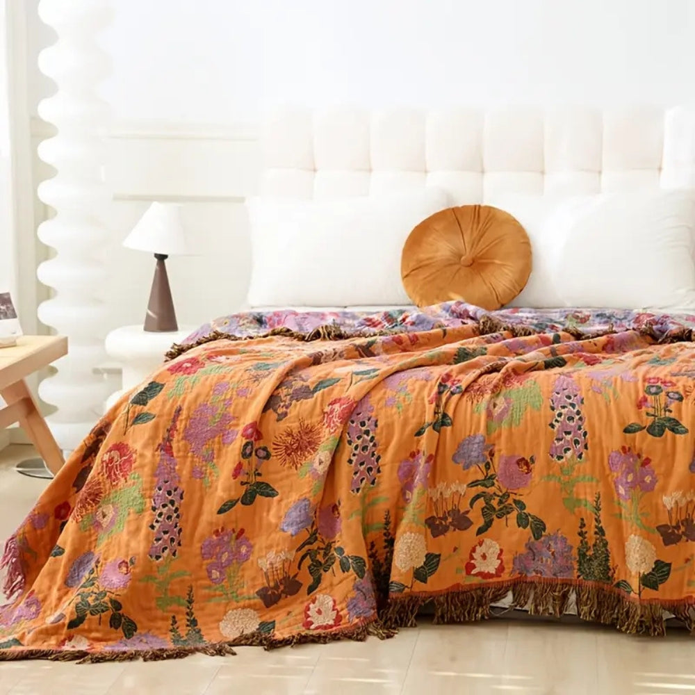 Orange cotton blanket with a vibrant floral pattern and tasseled edges draped over a bed.