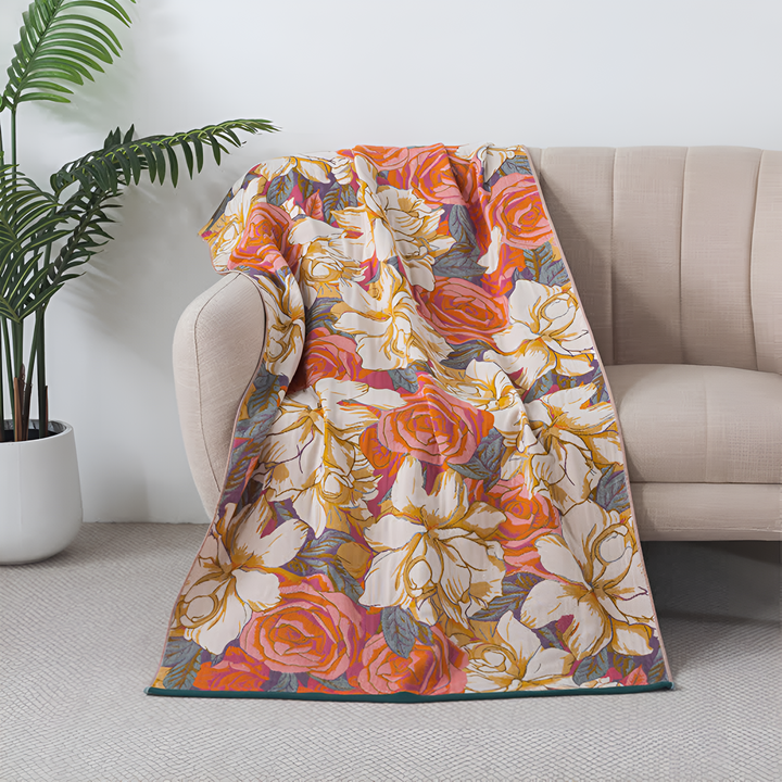 A plush throw blanket with a vibrant floral pattern featuring large roses in shades of coral, orange, pink, and cream, draped over a light-colored sofa.