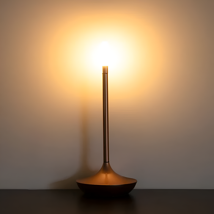 A modern copper table lamp with a wide base and a slender stem, emitting a warm, bright light against a neutral wall.