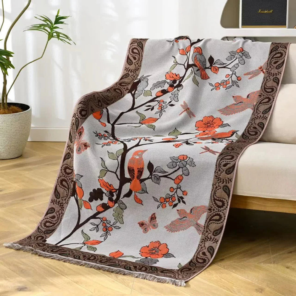 A light gray throw blanket with a brown paisley border and a pattern of birds, butterflies, dragonflies, and flowers.
