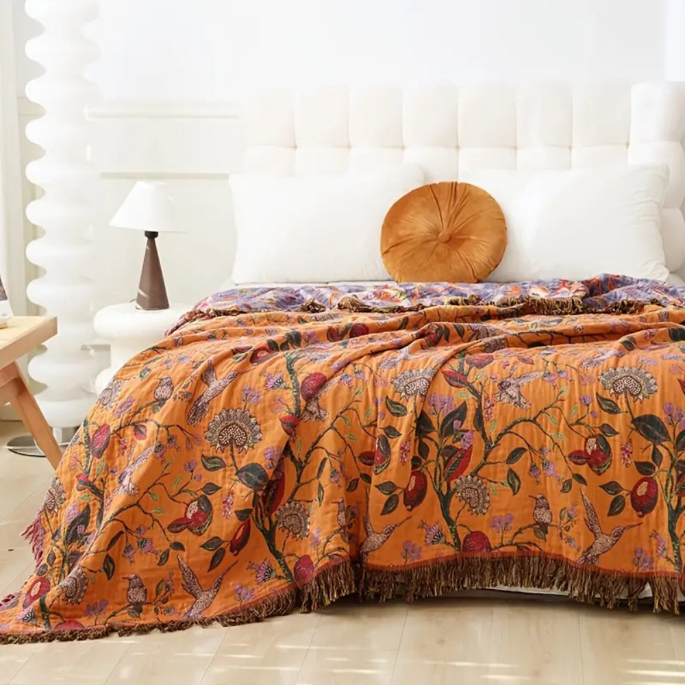 Orange cotton blanket with a floral and bird pattern, featuring a tasseled edge, draped over a bed.