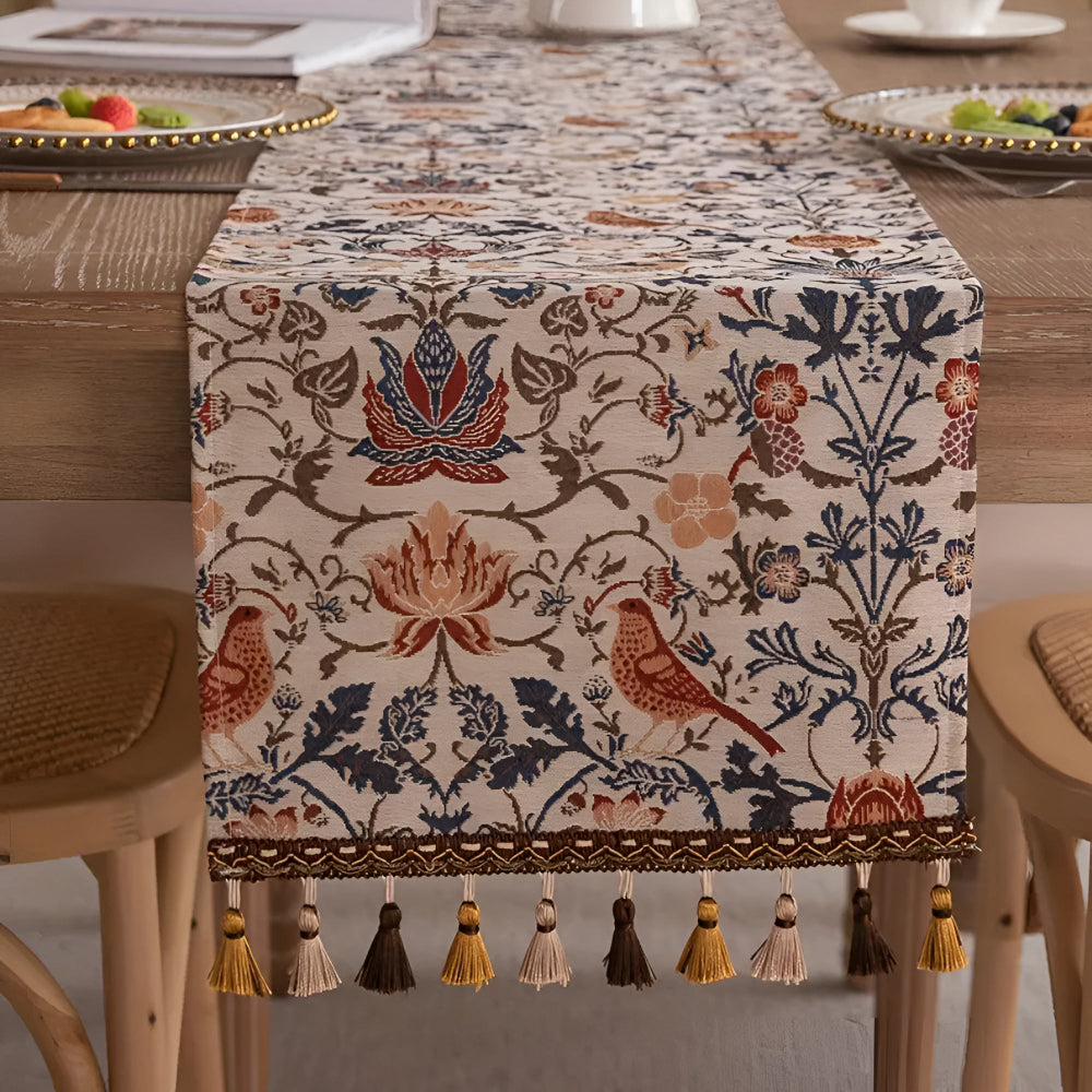 Vintage floral and bird jacquard table runner with tasseled edges on a wooden table.