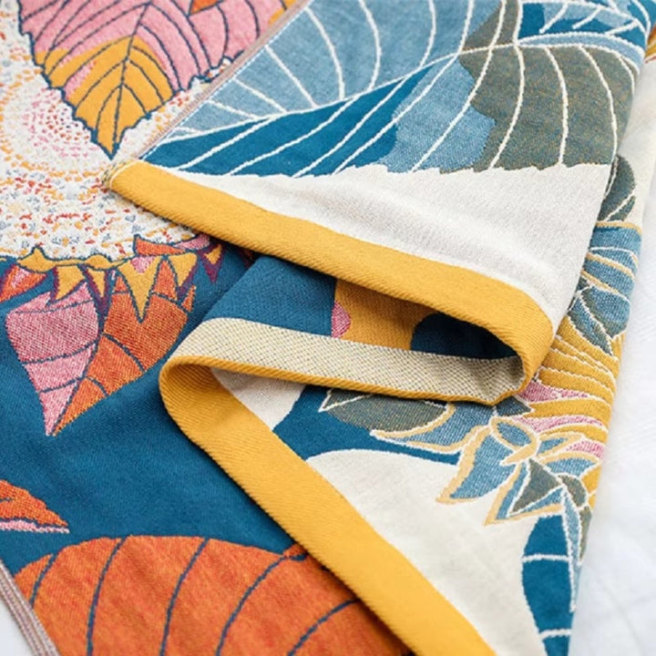 A folded cotton bath towel with a vibrant floral and leaf pattern in shades of blue, orange, pink, and yellow, featuring a yellow border.