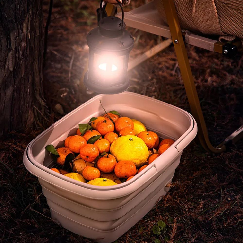 Collapsible storage basket filled with oranges and lemons, illuminated by a lantern.