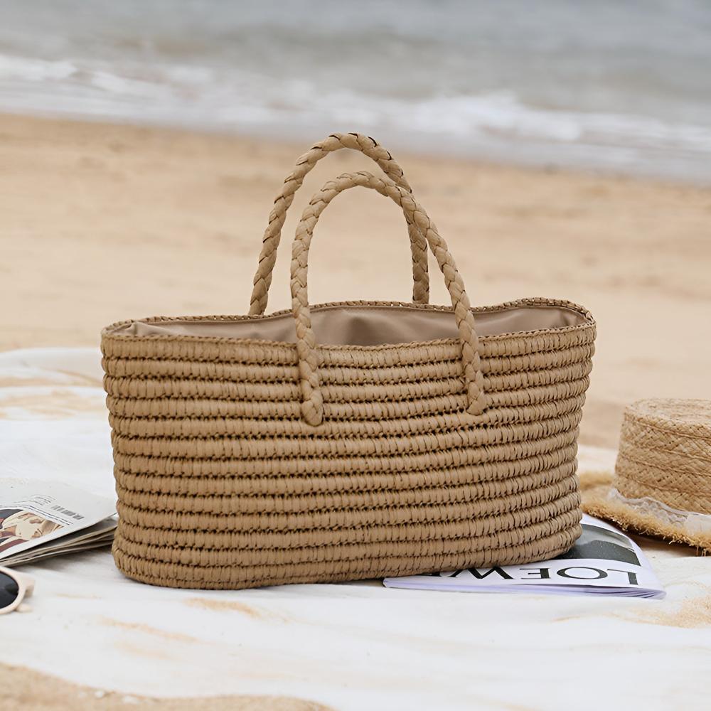 Handwoven straw tote bag with braided handles on a beach blanket with magazines and a straw hat.