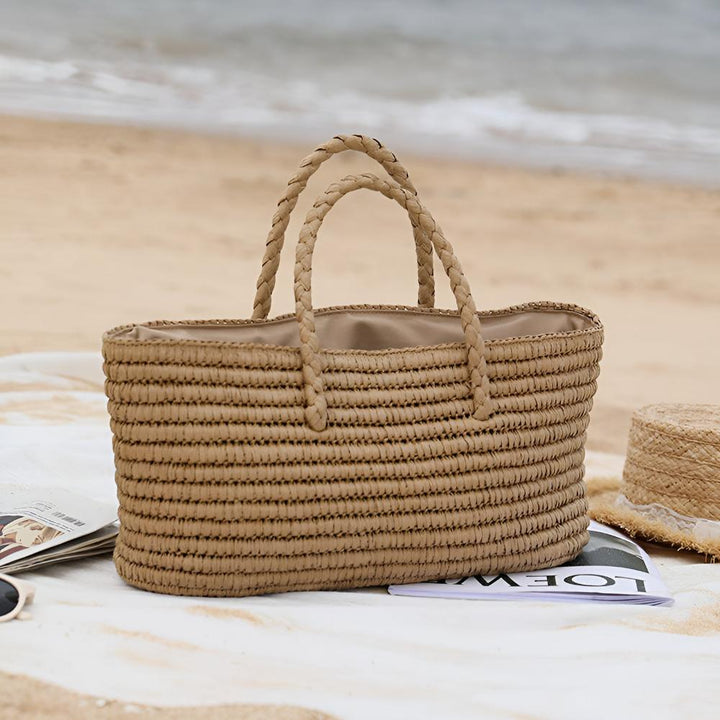 Handwoven straw tote bag with braided handles on a beach blanket with magazines and a straw hat.
