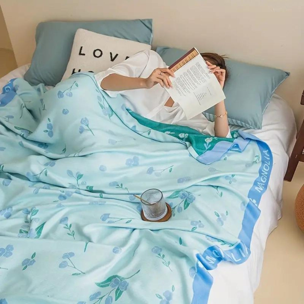 A person lies in bed reading under a light blue blanket with a floral pattern and a blue border.