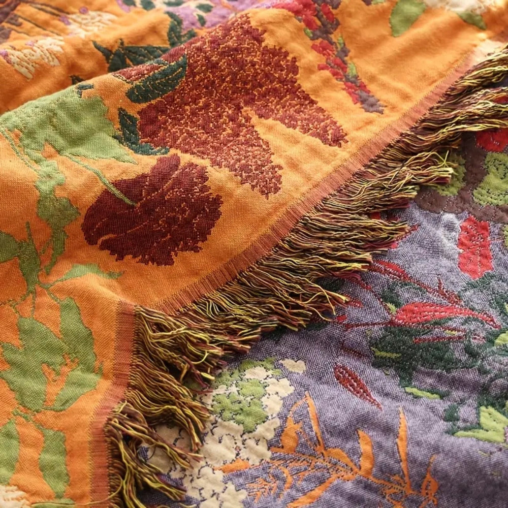A close-up of a woven cotton blanket with a floral pattern in orange, green, and purple, featuring a fringed edge.