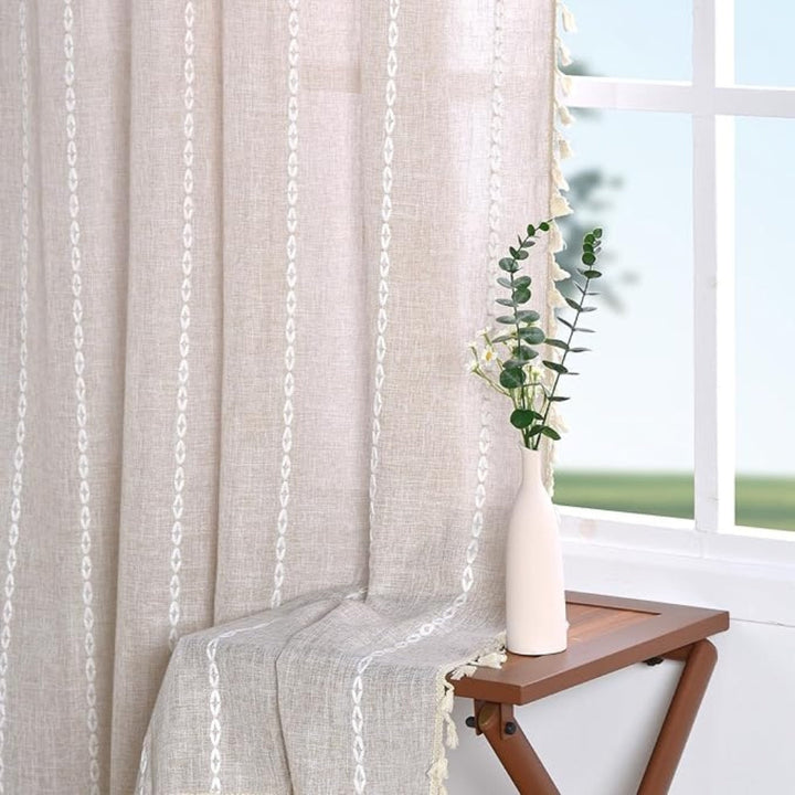 Beige boho cotton linen kitchen curtains with white embroidered stripes and tassels, draped over a small table with a vase of greenery.