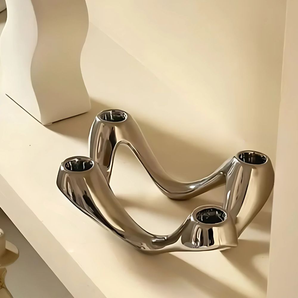 Sculptural, chrome-finished candle holder with four openings for candles, on a white shelf.