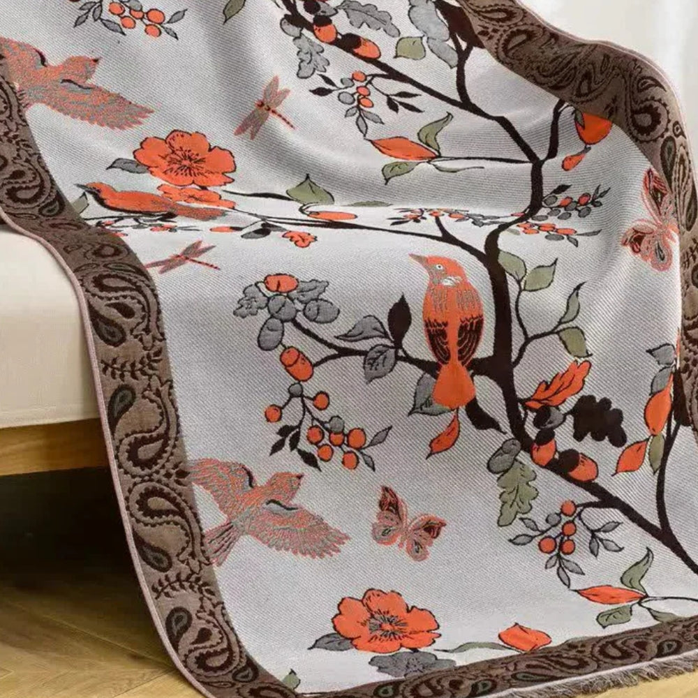 A light gray throw blanket with a brown paisley border, featuring a pattern of orange birds, butterflies, dragonflies, and floral branches.