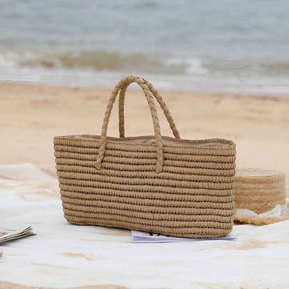 Handwoven straw tote bag with braided handles on a beach blanket with the ocean in the background.