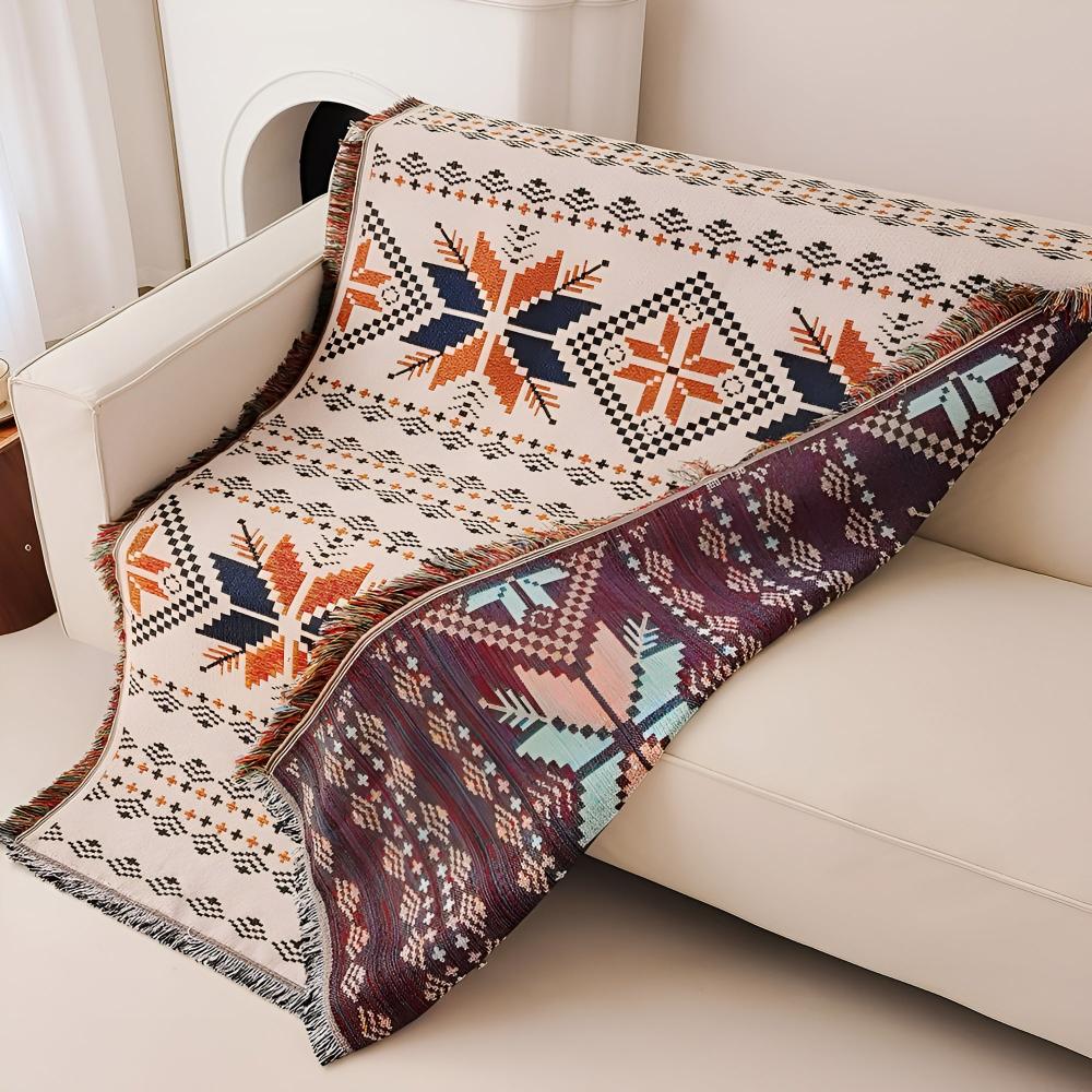 A cream-colored throw blanket with a geometric snowflake pattern in orange and navy, draped over a white sofa.