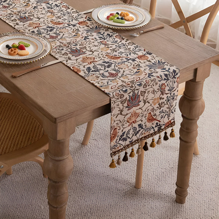 Vintage floral and bird jacquard table runner with tassels on a wooden table.