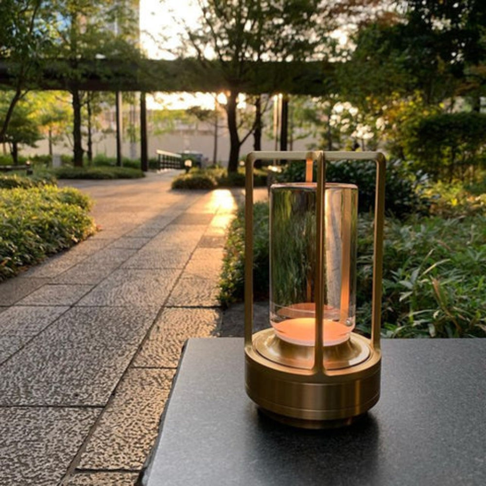 A brass and glass table lantern with a warm glow sits on a dark surface next to a stone pathway in a garden at sunset.