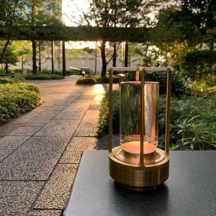 A brass and glass table lantern with a warm glow sits on a dark surface next to a stone pathway in a garden at sunset.