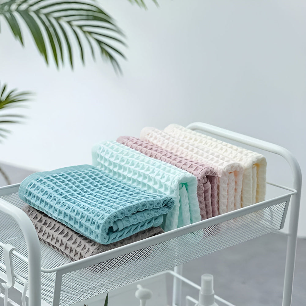 Four folded cotton waffle weave towels in pastel colors: blue, mint, pink, and cream, on a white mesh shelf.