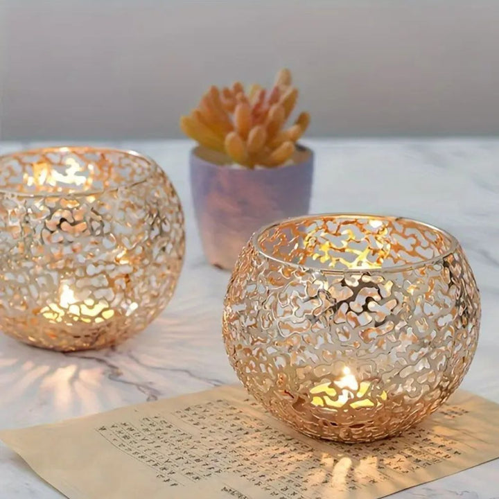 Two gold geometric iron tealight holders with intricate cutouts, one in front of a small potted succulent.