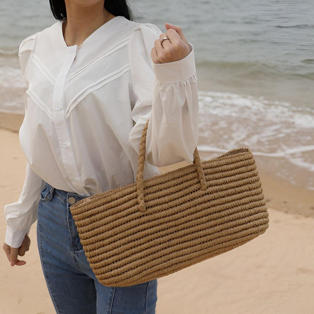 Woman on a beach holding a handwoven straw tote bag with braided handles.