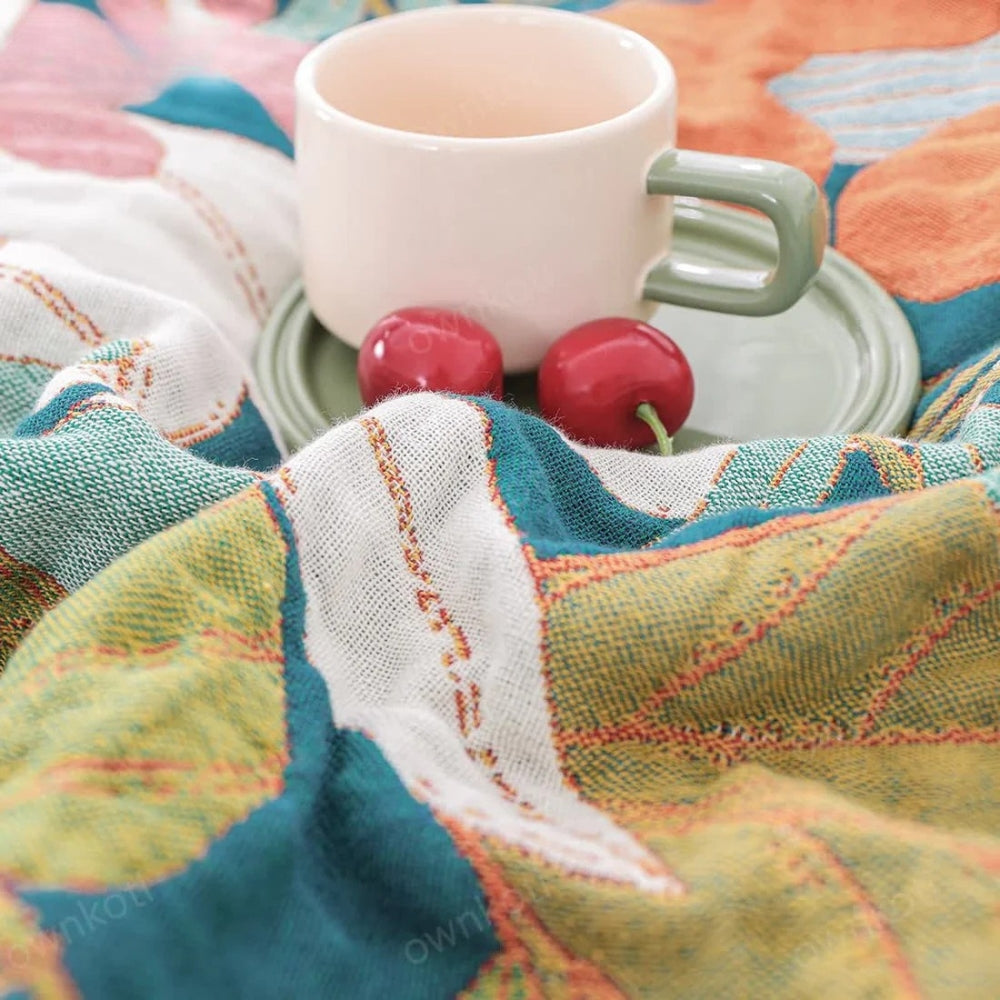 A cream mug with a green handle sits on a green saucer next to two red cherries on a colorful, textured gauze blanket.