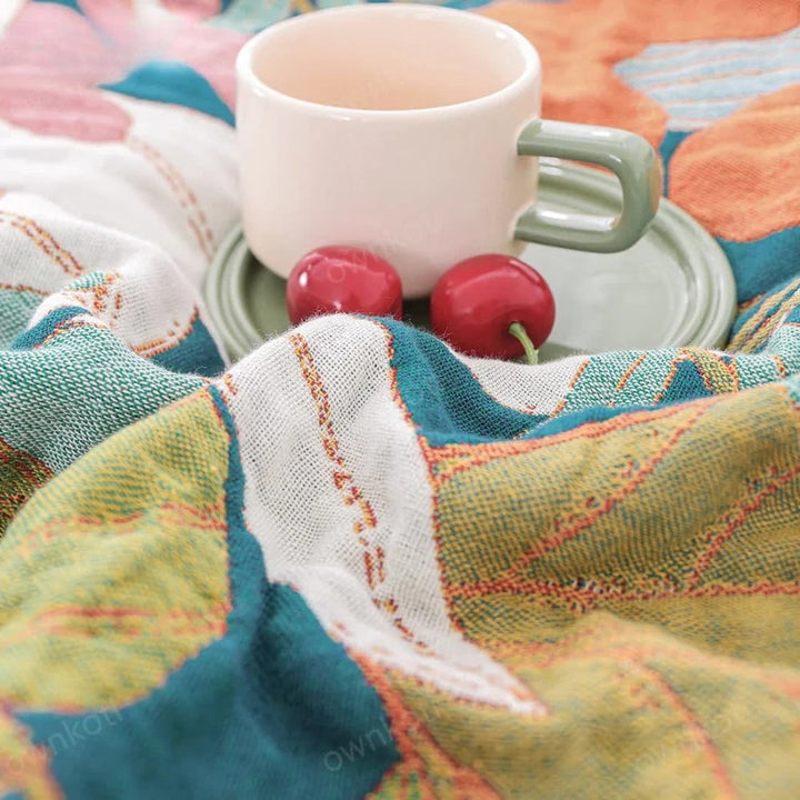 A cream mug with a green handle sits on a green saucer next to two red cherries on a colorful, textured gauze blanket.