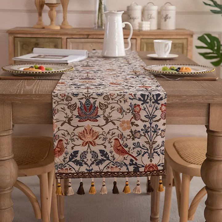 Vintage floral and bird jacquard table runner with tassels on a wooden table.