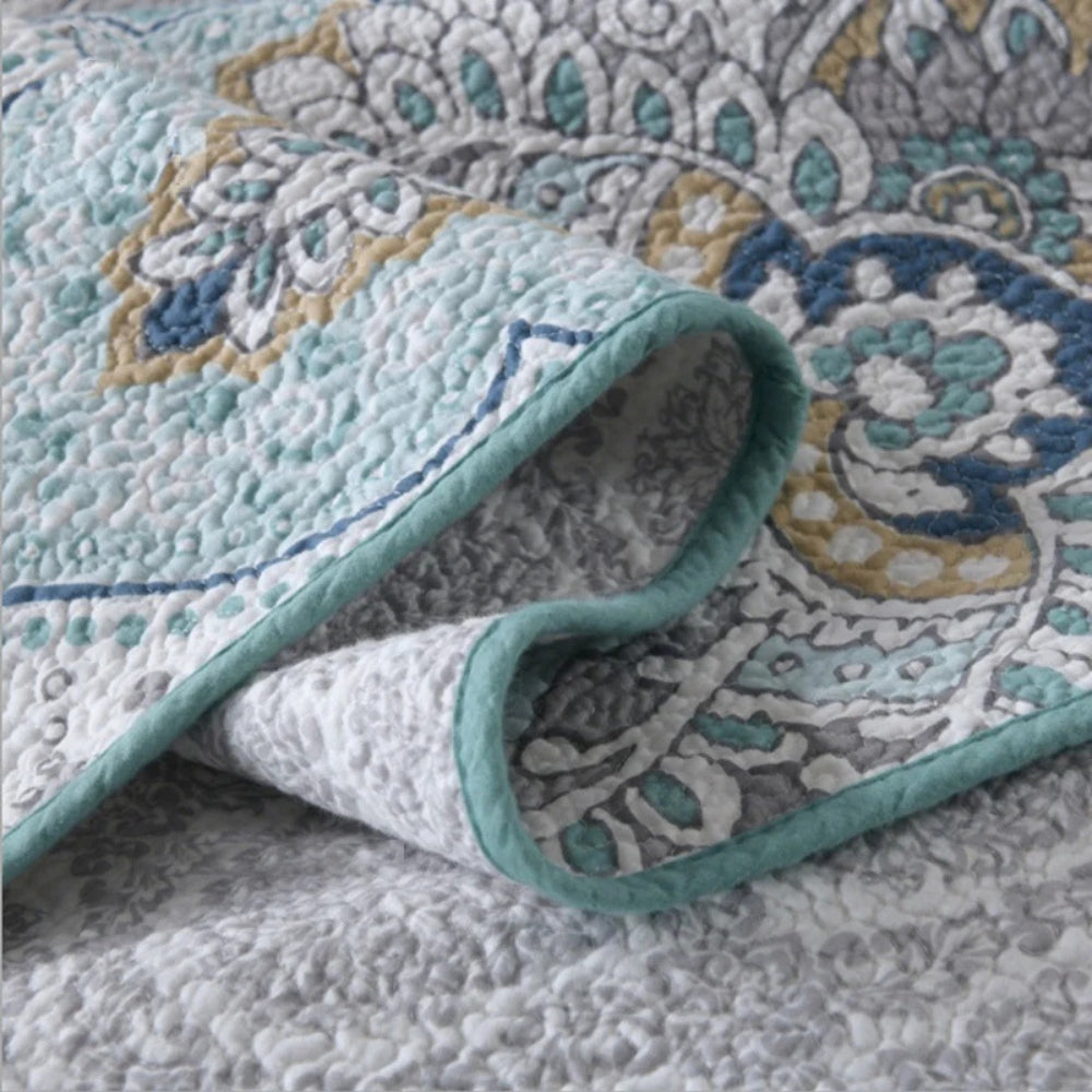 Close-up of a folded paisley aqua teal coverlet with a textured finish and blue, grey, and yellow floral patterns.
