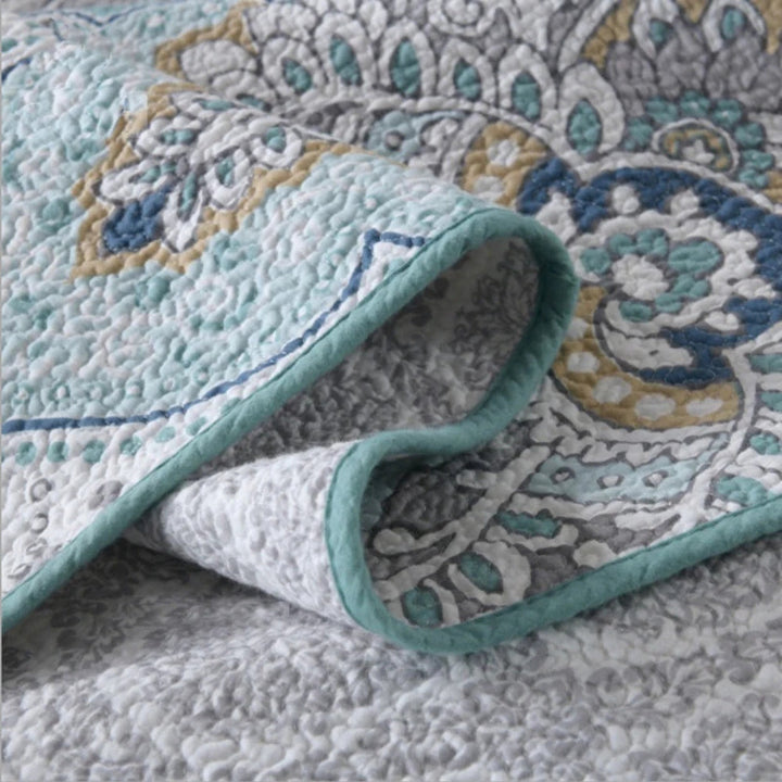 Close-up of a folded paisley aqua teal coverlet with a textured finish and blue, grey, and yellow floral patterns.