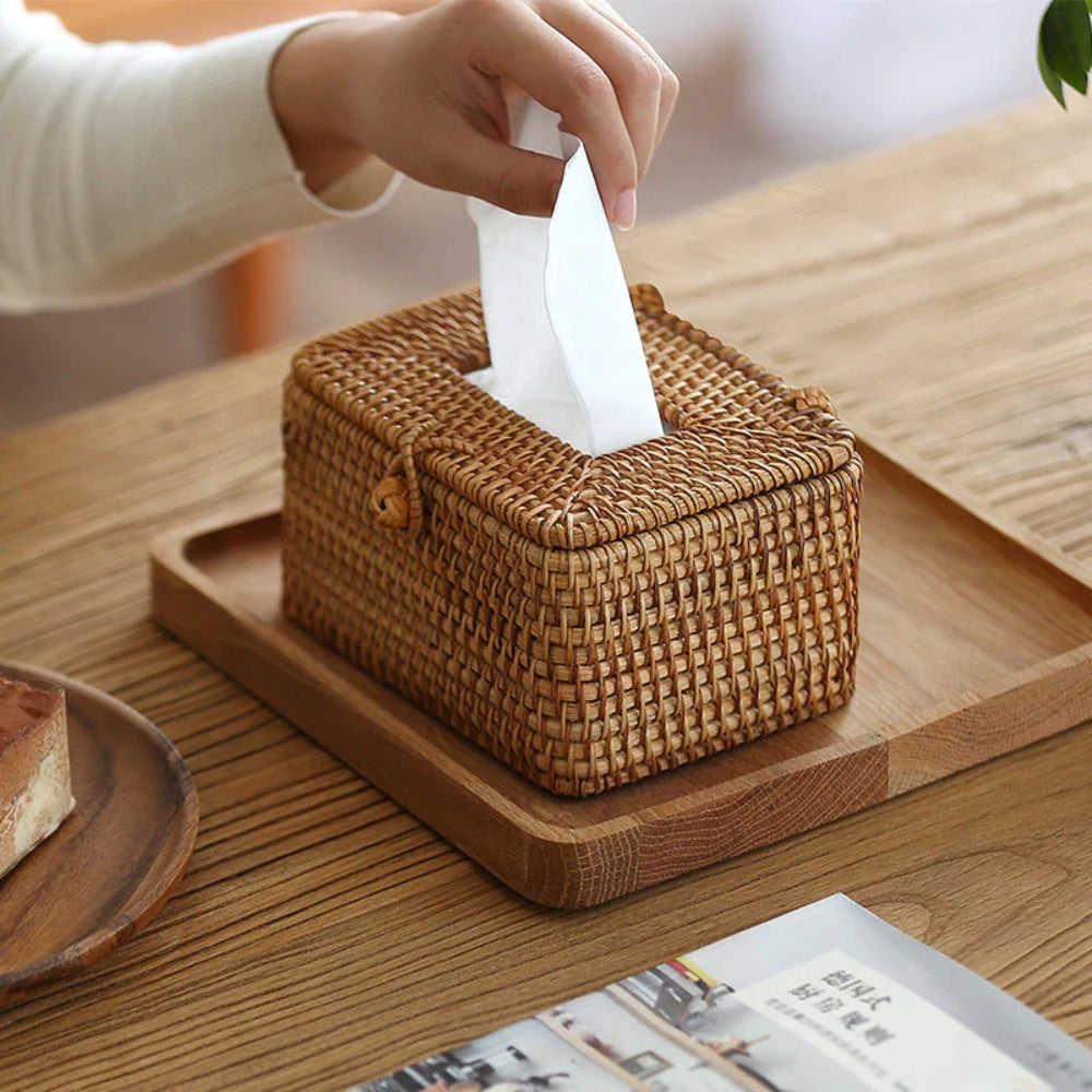Hand pulling a tissue from a handwoven rattan tissue box on a wooden tray.