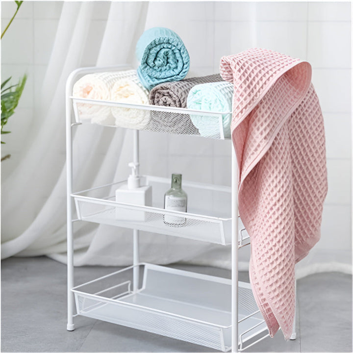 A white metal shelf unit holding rolled towels and toiletries, with a pink waffle-weave towel draped over the side.