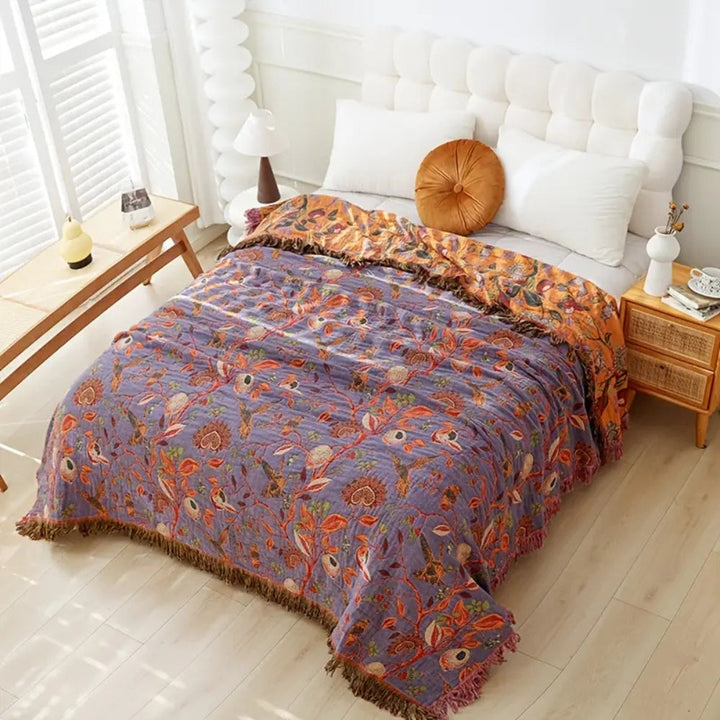 Botanic floral cotton tassel blanket in purple with orange accents and fringe, draped on a bed.