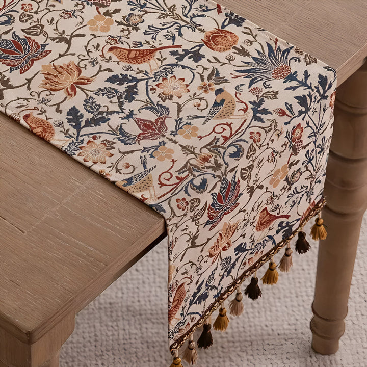 Vintage floral and bird jacquard table runner with tasseled ends on a wooden table.